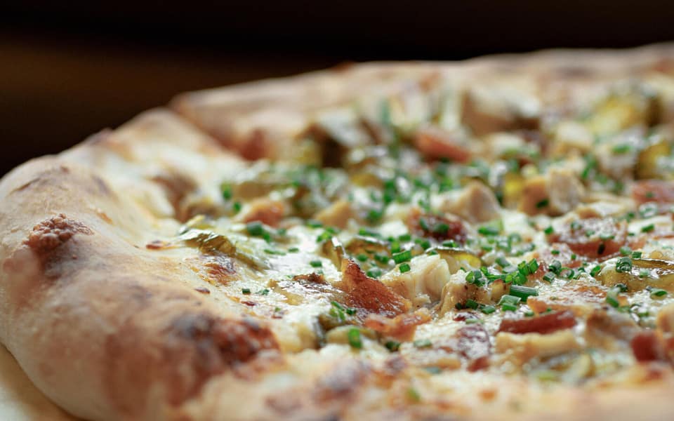 Close-up of a Pigzza pizza with melted cheese, smoked meat, pickled peppers, and chives on a blistered crust